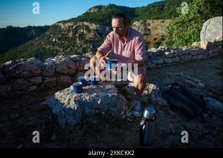 Homme prenant une pause pour profiter d'un café en plein air, entouré d'une vue panoramique sur la montagne. Détente et aventure dans la nature. Banque D'Images