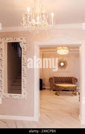 Un élégant couloir avec un lustre et un miroir orné sur un mur rose clair, menant à un salon élégant avec un canapé et un miroir décoratif. Banque D'Images