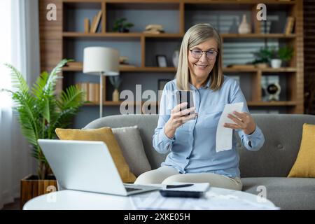 Femme senior assise sur le canapé à l'aide d'un smartphone et d'un ordinateur tout en vérifiant les reçus. Concept de travail à distance, gestion des finances à domicile, et accou en ligne Banque D'Images