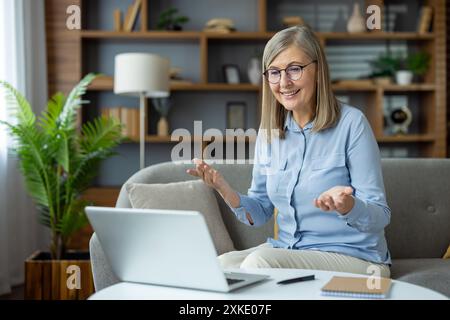 Femme senior chat vidéo sur ordinateur portable à la maison dans le salon lumineux. S'engager dans la communication virtuelle, détendue et confortable. Utilisation de la technologie à dai Banque D'Images