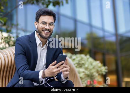 Jeune professionnel en tenue d'affaires assis sur le banc, souriant et écoutant de la musique sur smartphone avec des écouteurs. Style de vie urbain détendu dans un quartier d'affaires moderne capturé dans une photo vibrante. Banque D'Images