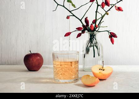 Pommes fraîches et verre de jus de pomme ou de cidre sur la table blanche. Vase avec des branches de rose musquée sur le fond. Récolte d'automne, concept de Thanksgiving. Banque D'Images