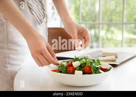 Jeune femme mettant des morceaux de fromage feta dans la salade à la maison Banque D'Images