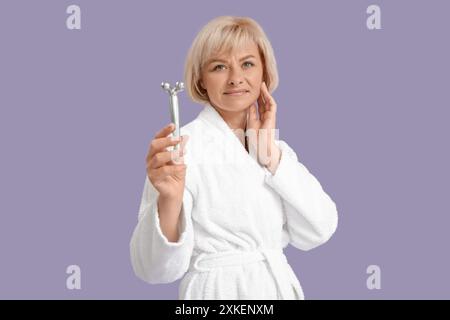 Belle femme heureuse mature en peignoir avec rouleau de massage facial sur fond violet Banque D'Images