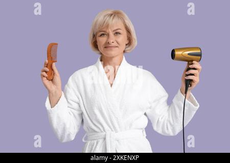 Belle femme heureuse mature en peignoir avec sèche-cheveux et peigne sur fond violet Banque D'Images