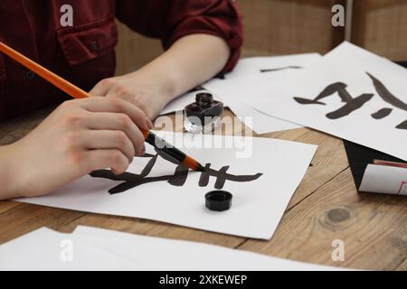 Calligraphie. Femme avec pinceau et encrier pratiquant l'écriture de hiéroglyphes chinois sur papier à la table en bois, gros plan Banque D'Images