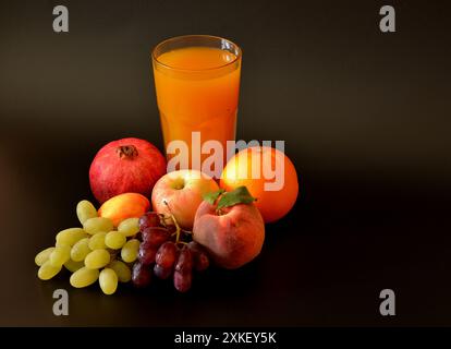 Un grand verre à facettes d'un mélange de jus de fruits sur un fond noir, entouré de raisins mûrs, pomme, pêche, grenade et orange. Gros plan. Banque D'Images