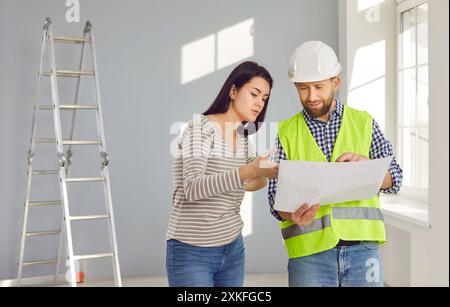 Discussion des détails des réparations et des changements dans la disposition de la maison. Rencontre jeune femme propriétaire et contremaître habillé en salopette et protecteur Banque D'Images