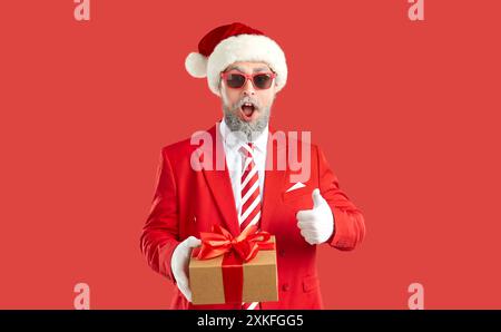 Portrait de drôle d'homme surpris qui tient le cadeau de Noël dans sa main et qui montre le pouce vers le haut. Banque D'Images