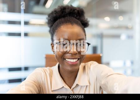 Femme souriante portant des lunettes prenant selfie dans un bureau moderne pendant un appel vidéo. Capture les émotions positives, le cadre professionnel et la communication virtuelle. Banque D'Images