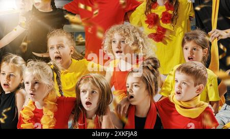 Groupe d'enfants vêtus de tenues rouges, jaunes et noires éclatantes regardant leur équipe préférée jouer sur le terrain, et des confettis dorés tombant Banque D'Images