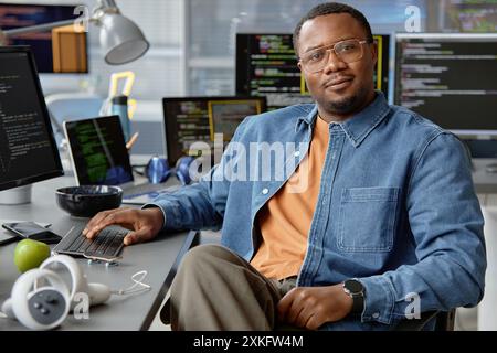 Portrait d'un développeur de VR afro-américain masculin confiant posant alors qu'il était assis au bureau de travail regardant la caméra dans le bureau du programmeur informatique Banque D'Images