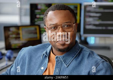 Gros plan portrait du jeune ingénieur logiciel de VR masculin afro-américain dans des lunettes joyeusement souriant regardant la caméra tout en étant assis dans le bureau de programmation informatique, espace de copie Banque D'Images