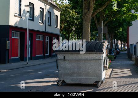 Une grande benne à ordures en métal se trouve sur le trottoir à côté d'une rangée de magasins par une journée ensoleillée. Banque D'Images