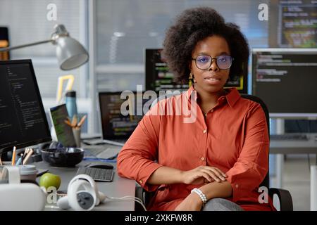 Portrait moyen d'une femme afro-américaine bouclée révélatrice VR portant des lunettes et une chemise orange regardant la caméra tout en étant assis au bureau de travail avec plusieurs écrans d'ordinateur, espace de copie Banque D'Images