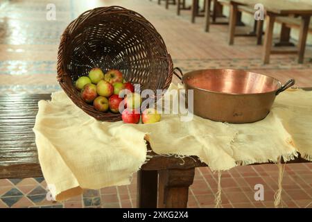 Nature morte faite avec un panier en osier rempli de pommes et un bassin en cuivre tous les deux sur le dessus d'une toile de ligne étalée sur une table. Banque D'Images