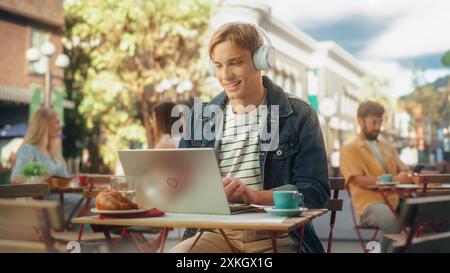 Freelance portant des écouteurs blancs et utilisant un ordinateur portable pour le travail en ligne à distance dans la ville. Jeune homme créatif prenant le petit déjeuner et le télétravail d'un café sur une journée d'été ensoleillée Banque D'Images