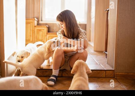 Une petite fille est assise sur les marches dans une pièce et caresser les chiots labrador. Un enfant mignon s'amuse avec des chiens à la maison. Un enfant souriant prend CA Banque D'Images