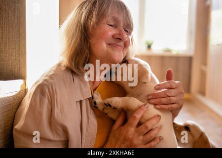 Une femme mature dans des vêtements décontractés embrasse un chiot à la maison. Un animal de compagnie lèche le visage de son propriétaire. Un retraité s'occupe d'un petit Golden retriever. Un âge moyen w Banque D'Images