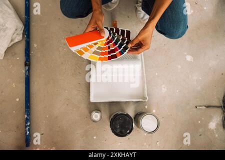 Gros plan des mains féminines tenant la palette de couleurs, les boîtes et le bac à peinture sur le sol en béton. La jeune femme choisit la couleur de la peinture en utilisant des échantillons de peinture. À Banque D'Images