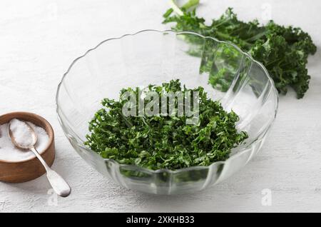 Feuilles de chou frisé hachées, écrasées avec du sel dans un bol transparent sur un fond gris clair. Des aliments détox sains. Banque D'Images