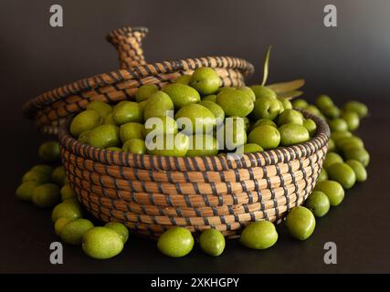 Olives vertes dans un panier en osier avec couvercle. Il y a des olives autour du panier. Fond noir. Banque D'Images