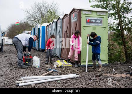 Femme dans Red Workman installant des toilettes et des sanitaires , surveillée par des enfants migrants curieux, à destination du Royaume-Uni. Dunkerque, France. Duinkerken la Jungle Nord pas de Calais France Copyright : xGuidoxKoppesxPhotox Banque D'Images