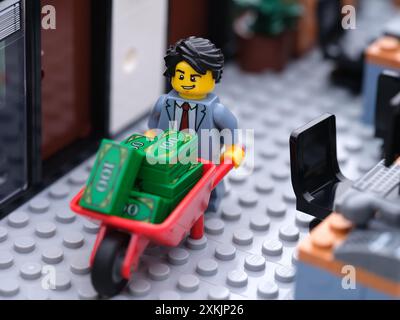 Tambov, Fédération de Russie - 19 juillet 2024 Un homme d'affaires Lego minifigure marchant dans un bureau et poussant une brouette pleine d'argent. Banque D'Images