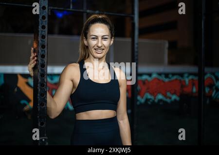 Une femme portant un soutien-gorge de sport noir et des leggings s'appuie contre un support de gymnastique en métal. Le fond est flou, et il y a des graffitis sur le mur. Banque D'Images