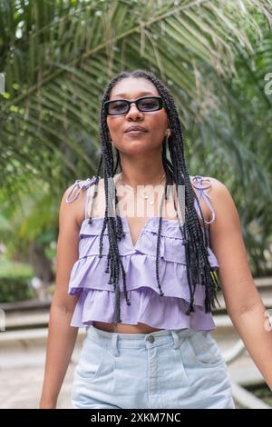 Femme confiante dans un haut violet et lunettes de soleil debout sous les palmiers Banque D'Images