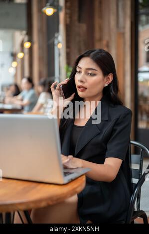 Une femme d'affaires asiatique confiante et attrayante dans un costume noir parle au téléphone avec son client tout en travaillant sur son ordinateur portable dans un café Banque D'Images