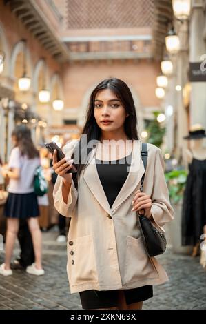 Une femme asiatique attrayante et confiante dans des vêtements à la mode se tient dans une rue commerçante de la ville, tenant son smartphone et regardant l'appareil photo. Banque D'Images