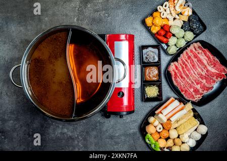 Une casserole rouge avec un couvercle se trouve sur un comptoir à côté d'un brûleur rouge. Une variété d'aliments sont sur une table, y compris de la viande, des légumes et un bol de soupe. C Banque D'Images