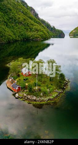 Une cabane rouge se trouve sur une petite île verte entourée d'un fjord calme et clair en Norvège. L'île est couverte d'une végétation luxuriante et est nichée entre deux collines boisées. Lovrafjorden, Norvège Banque D'Images