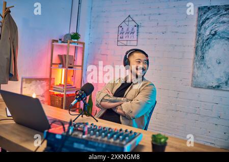 Un homme asiatique sourit avec éclat tout en portant des écouteurs et en enregistrant dans son studio de podcast. Banque D'Images