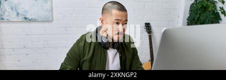 Un bel homme asiatique, portant des écouteurs, se concentre intensément sur un écran d'ordinateur dans son studio, entouré d'instruments. Banque D'Images