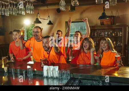 Les fans enthousiastes vêtus de chemises orange s'assoient au bar avec des verres de bière, regardant des événements sportifs et montrant des expressions de célébration. Banque D'Images