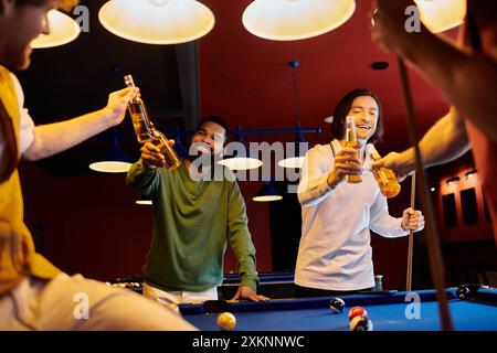 Un groupe d'amis porte un toast avec des bouteilles de bière tout en jouant au billard dans une pièce faiblement éclairée. Banque D'Images