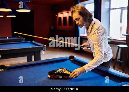 Un homme en chemise blanche décontractée organise une partie de billard dans une salle de billard faiblement éclairée. Banque D'Images