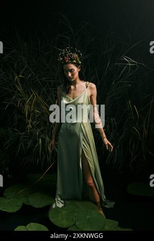 Une femme se tient gracieusement sur des nénuphars dans un marais, portant une robe verte fluide et une couronne florale. Banque D'Images