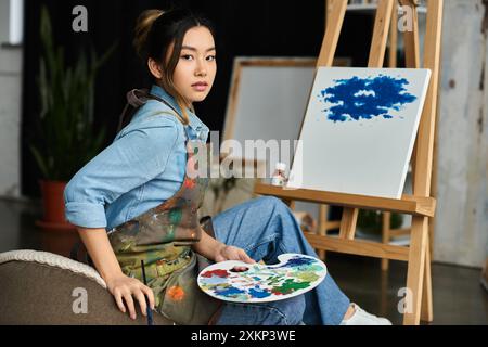 Une artiste asiatique, vêtue d’une chemise en Jean et d’un tablier, s’assoit dans son atelier avec une palette de peinture dans ses mains, prenant une pause de son propre toit Banque D'Images