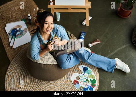 Une jeune femme asiatique, vêtue d'un tablier, s'assoit confortablement sur une chaise de sac de haricots dans son atelier d'art, tenant un pinceau et souriant. Banque D'Images