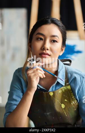Une jeune artiste asiatique, portant un tablier enduit de peinture, tient un pinceau dans son atelier. Banque D'Images