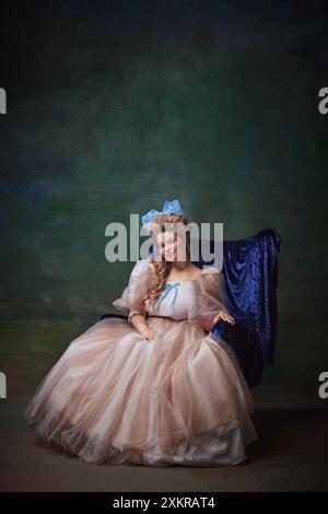 Portrait de jeune femme élégante dans une robe d'époque luxueuse assise sur une chaise bleue sur fond vert vintage. personne royale médiévale Banque D'Images