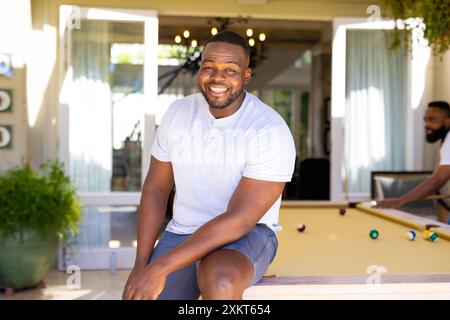 Homme souriant assis sur la table de billard, profitant du temps libre avec des amis à la maison Banque D'Images