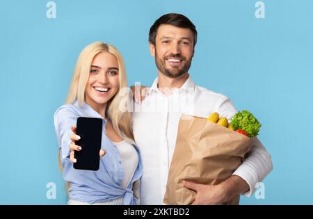 Une femme souriante tient un smartphone avec un écran noir, tandis qu'un homme à côté d'elle tient un sac en papier rempli de provisions. Ils regardent tous les deux directement Banque D'Images