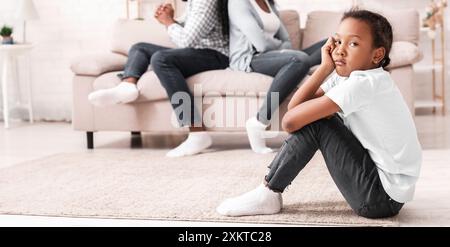 Concept d'arguments familiaux. Triste petite fille afro-américaine assise séparément des parents après leur querelle, focalisation sélective sur l'enfant Banque D'Images