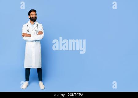 Un médecin indien barbu, portant une blouse de laboratoire blanche, se tient debout les bras croisés sur un fond bleu. Il regarde l'espace de copie, avec un sli Banque D'Images