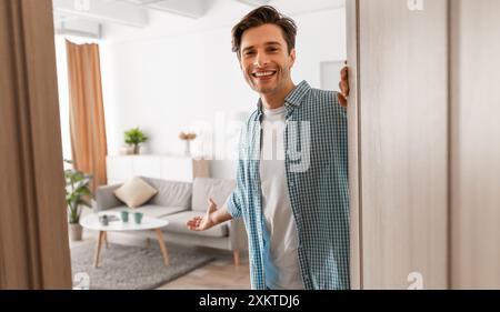 Portrait de l'homme joyeux accueillant visiteur invitant à entrer dans sa maison, jeune homme heureux debout dans la porte de l'appartement moderne, holdin masculin millénaire Banque D'Images