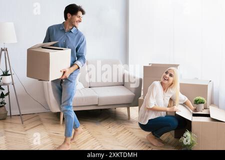 Un jeune homme porte une boîte dans ses bras et sourit à une jeune femme assise sur le sol déballant des boîtes dans un nouvel appartement. Banque D'Images